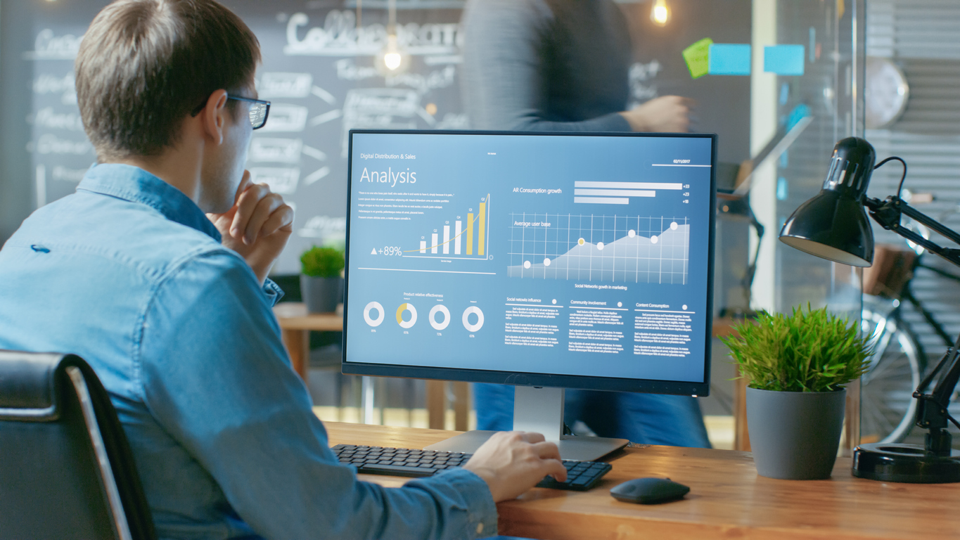 Analyst Works On A Personal Computer Showing Statistics, Graphs And Charts. In The Background His Coworker And Creative Office.